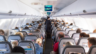  Innenansicht von einem Flugzeug mit sitzenden Passagieren und einer bedienenden Flugbegleiterin während eines Fluges.