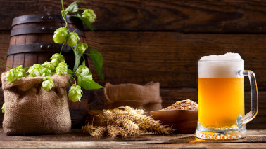 Ein gefülltes Bierglas steht vor einem Arrangement aus einem Holzfass, mehreren Leinensäcken und einer Holzschale mit Getreide. Dazwischen liegen Hopfendolden und Weizenähren.