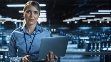 IT-Spezialistin steht mit geöffnetem Laptop in der Hand in einem großen Raum mit mehreren Servern.