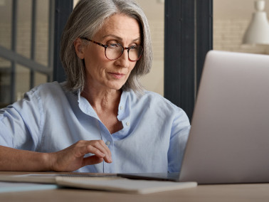 Eine ältere Frau mit Brille sitzt in lockerer Haltung an einem Tisch mit Laptop in einem modernen Arbeitsraum.