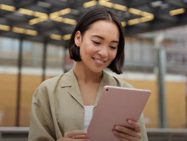 Eine junge dunkelhaarige junge Frau in Business-Kleidung schaut leicht lächelnd auf ein Tablet in ihrer Hand. Im Hintergrund ist eine moderne, helle Lagerhalle zu erkennen.