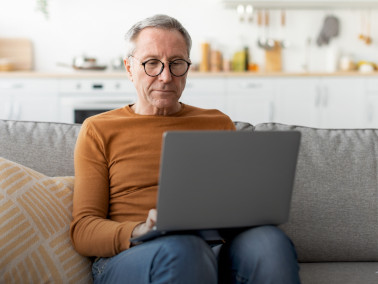 Ein älterer Mann sitzt mit einem Laptop auf den Knien auf einer Couch. Er leht sich leicht an ein Kissen und neben ihm steht eine Tasse, im Hintergrund ist unscharf eine helle Küche zu sehen.