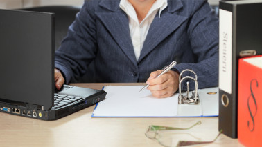 Eine Frau sitzt an einem Schreibtisch und schreibt auf einem Blatt in einem Ordner, während sie etwas in ihren Laptop tippt.
