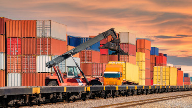 Containerlader transportiert von einem Güterzug Container auf eine Sattelzugmaschine, gespaltete Container befinden sich im Hintergrund.