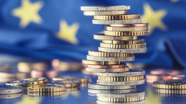 Münzenstapel stehen auf einem Tisch, im Hintergrund sieht man unscharf eine Europaflagge.