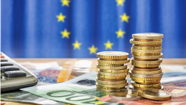 Geldscheine und Münzenstapel liegen bzw. stehen auf einem Tisch, daneben sieht man eine Ecke eines  Taschenrechners. Im Hintergrund sieht man unscharf eine Europaflagge.