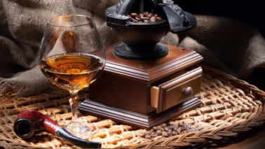 Eine alte, mit Kaffeebohnen gefüllte, Kaffeemühle und ein Glas Cognac stehen auf einem (Rattan)-Tablett, davor liegt eine Pfeife.