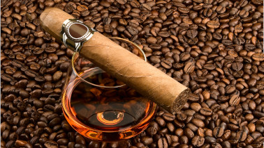Eine Zigarre liegt auf einem Glas Cognac, welches auf einer Fläche mit Kaffeebohnen steht.