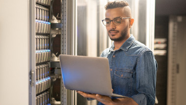 Ernsthafter qualifizierter junger IT-Spezialist mit Bart, der vor einem modernen Server steht und einen Laptop verwendet, während er Netzwerkprozesse überwacht.