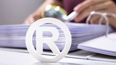 Das "Registered Trademark"-Zeichen (R in einem Kreis) steht ausgeschnitten vor einem geöffneten Ordner, in dem eine Person blättert.