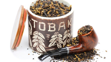Mit Tabak gefüllte Pfeife und Tabakdose gefüllt mit Tabak (Aufschrift "TOBAK"). Deckel der Tabakdose lehnt an Dose.
