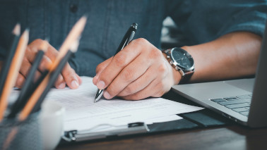 Person schreibt mit einem Kugelschreiber auf einem Papier, neben ihm steht ein aufgeklappter Laptop. Person schreibt mit einem Kugelschreiber auf einem Papier, neben ihm steht ein aufgeklappter Laptop.