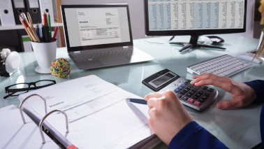 Eine Person tippt auf einem Taschenrechner und zeigt mit einem Stift auf Unterlagen in einem Ordner, im Hintergrund steht auf dem Schreibtisch ein Laptop und ein Computer.