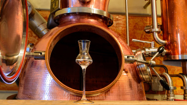 Ein mit Schnaps gefülltes Glas steht in einer Schnapsproduktion vor einem geöffneten Brennkessel